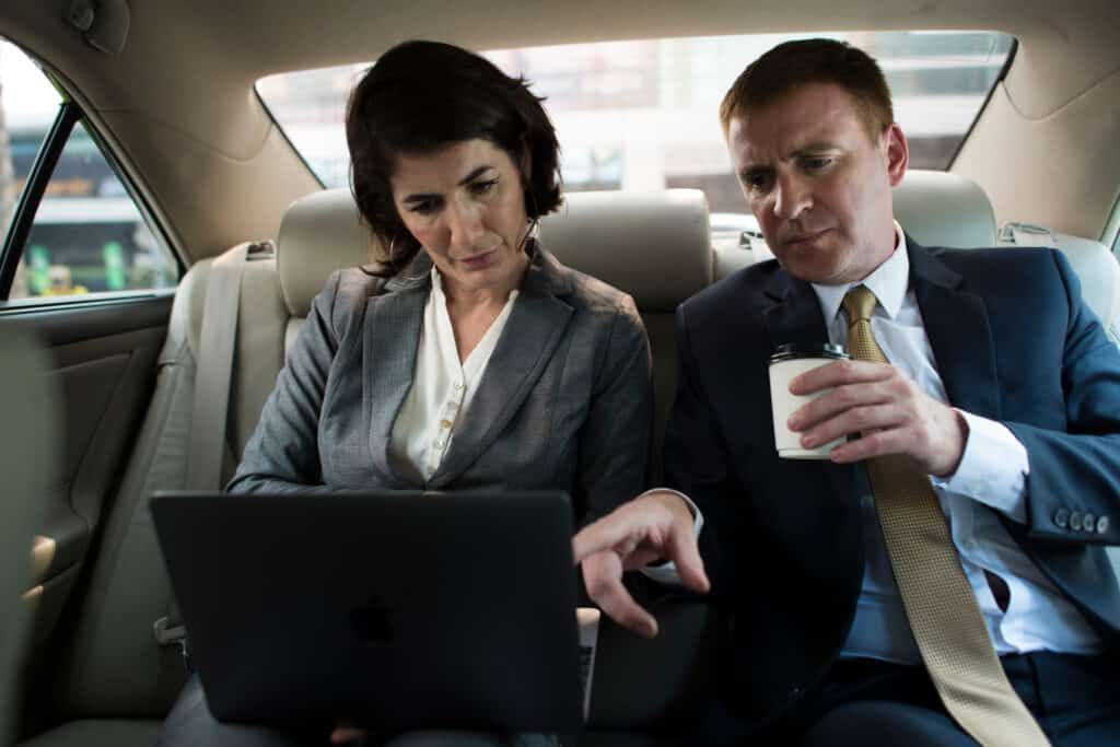 Two businesspeople, a woman and a man, working on a laptop in a car.
