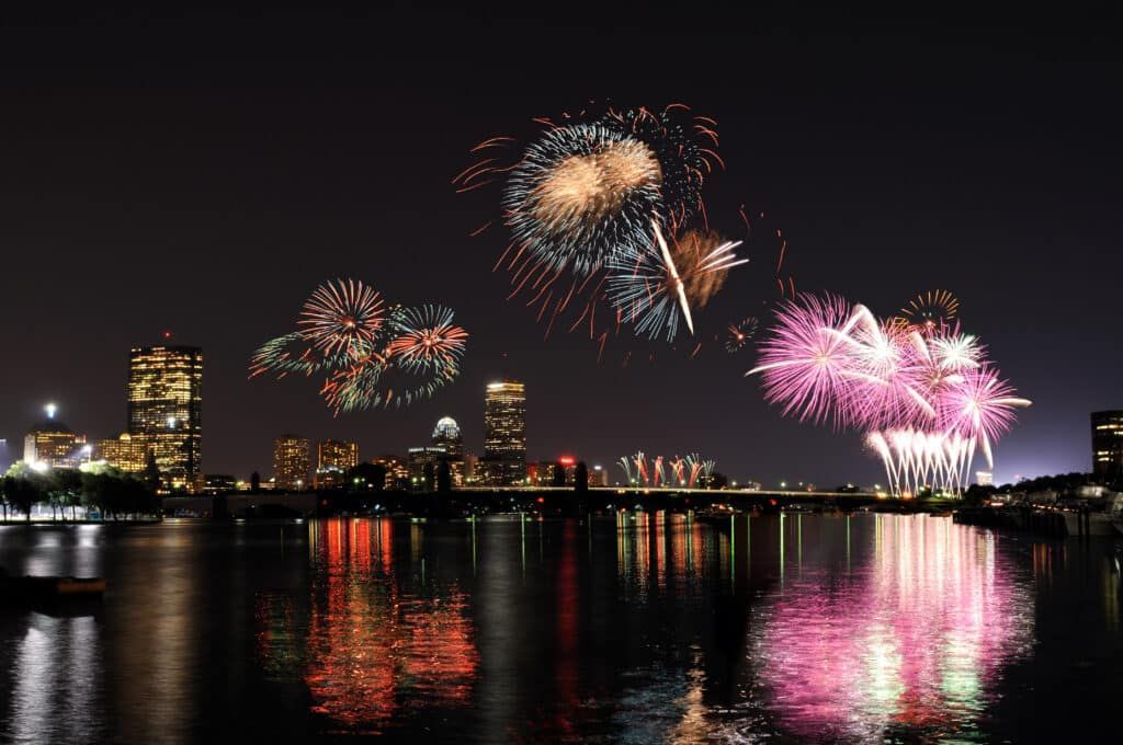 Fireworks explode over Boston skyline, reflecting in the water at night.