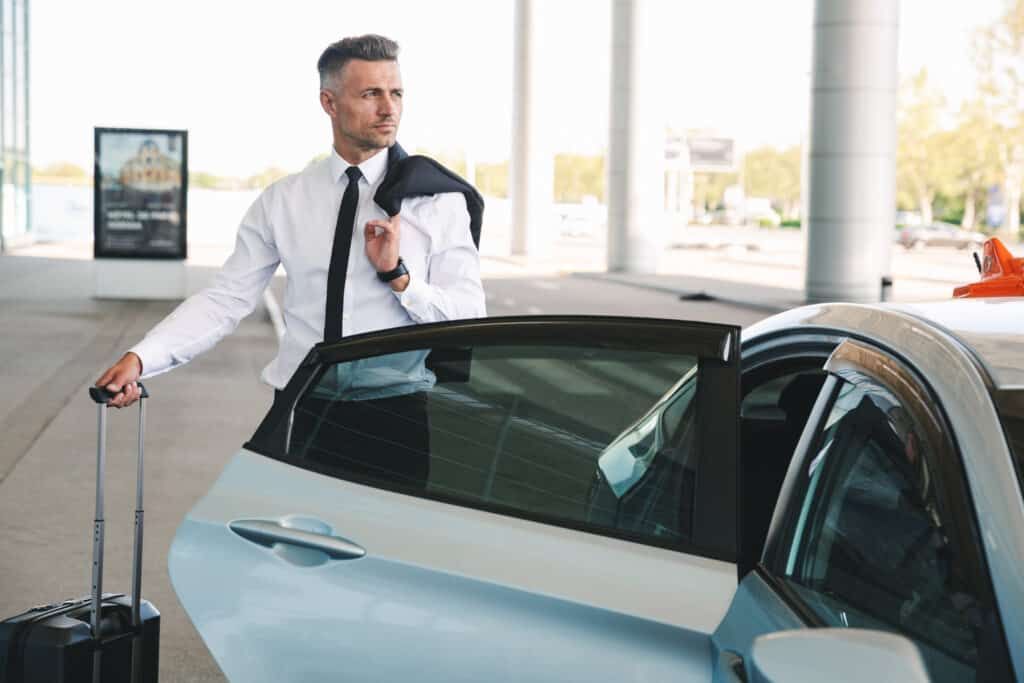 Businessman with luggage, exiting a blue car near a building.