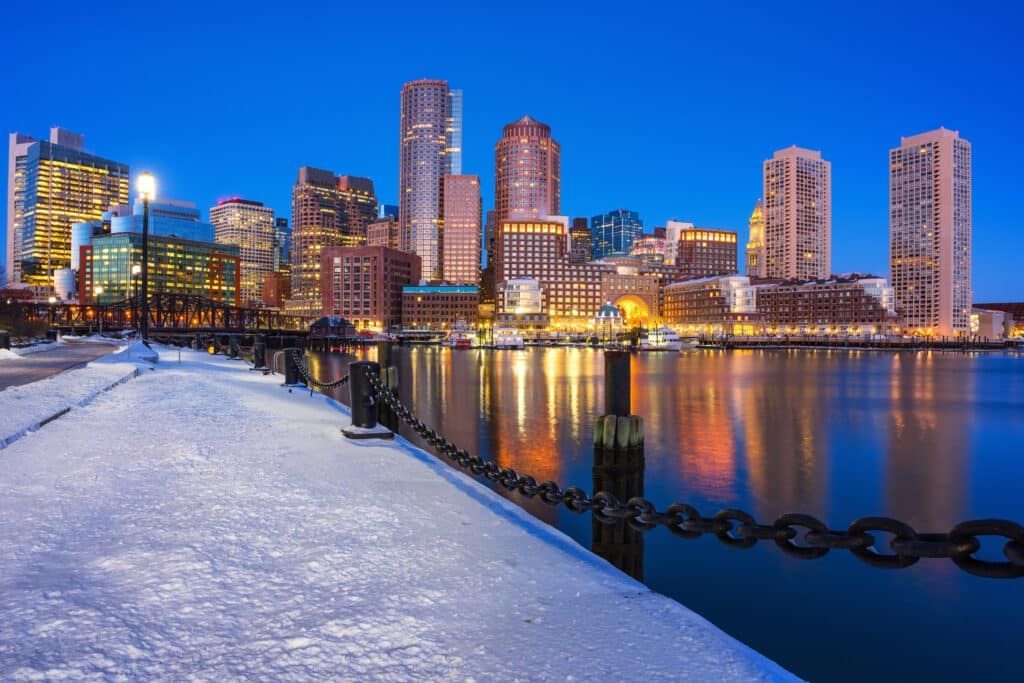 Boston skyline at dusk, reflecting in the calm water. Snow-covered pier in foreground, buildings aglow.