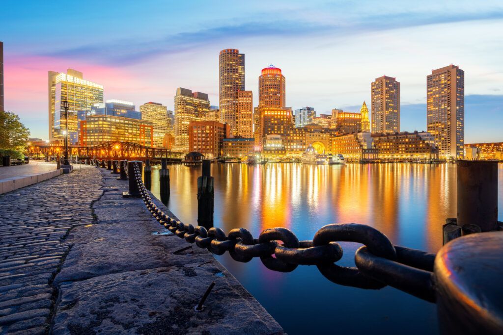 Boston skyline at dusk, illuminated buildings reflected in calm water, foreground of stone pier with chain.