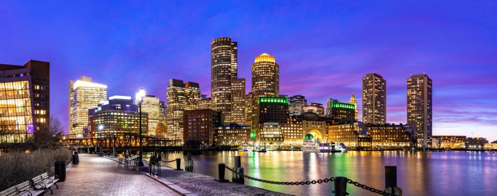 Boston skyline at dusk, buildings lit up, waterfront view.