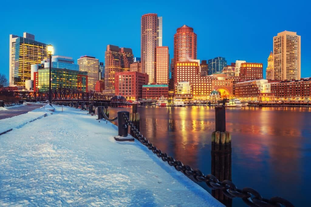 Boston skyline at dusk, overlooking water and a snow-covered pier. Buildings illuminated with lights.