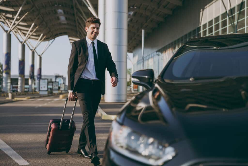 Man in suit with a suitcase walks toward a black car outside a building.