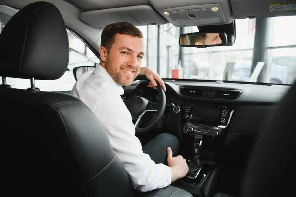 Smiling man sitting in a car, looking over shoulder. White shirt, black interior. Dealership setting.