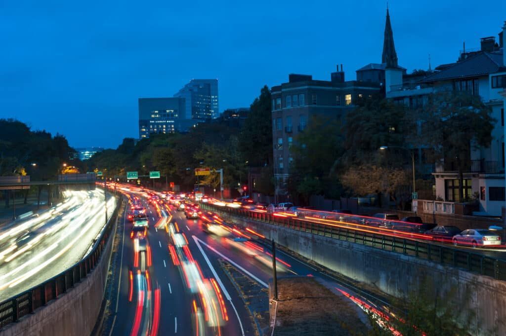 Nighttime view of a highway with long streaks of car lights, buildings in the background under a blue sky.