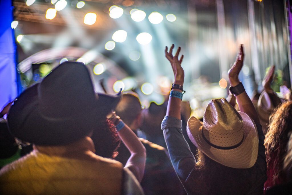 People in a crowd at a concert, raising hands, wearing cowboy hats, stage lights in the background.