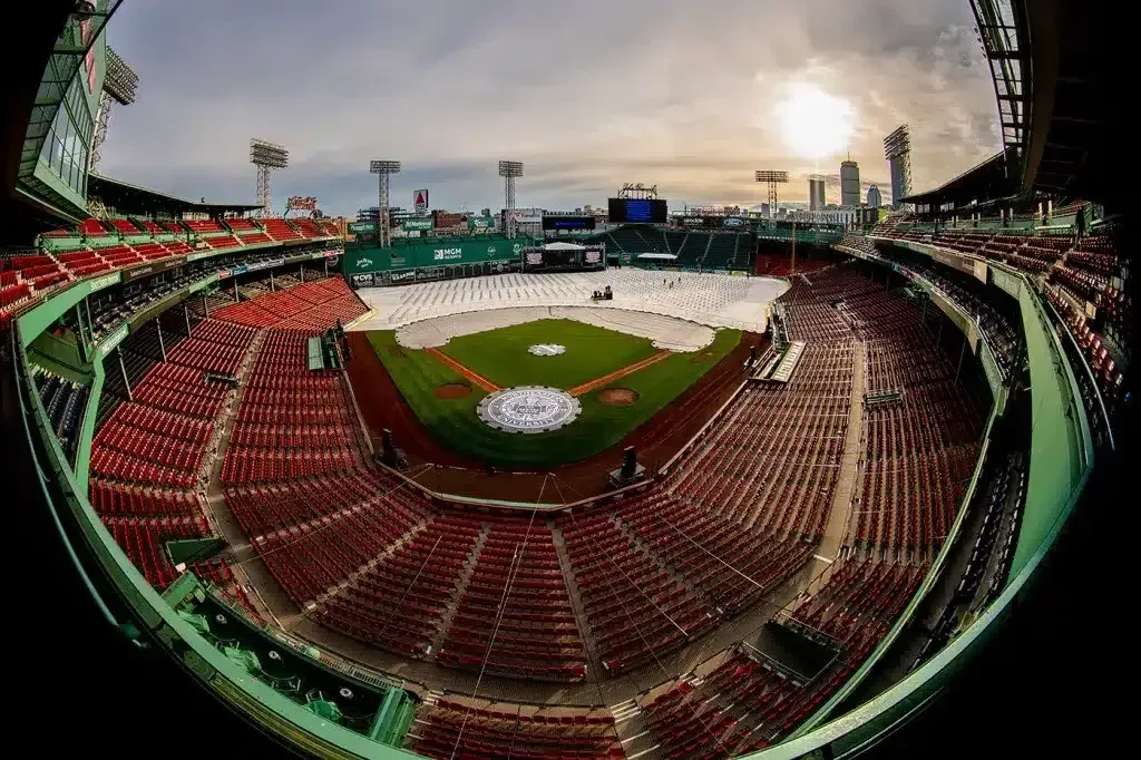 Empty Fenway Park from a wide angle; red seats, green field covered by white tarp, cityscape in the distance.