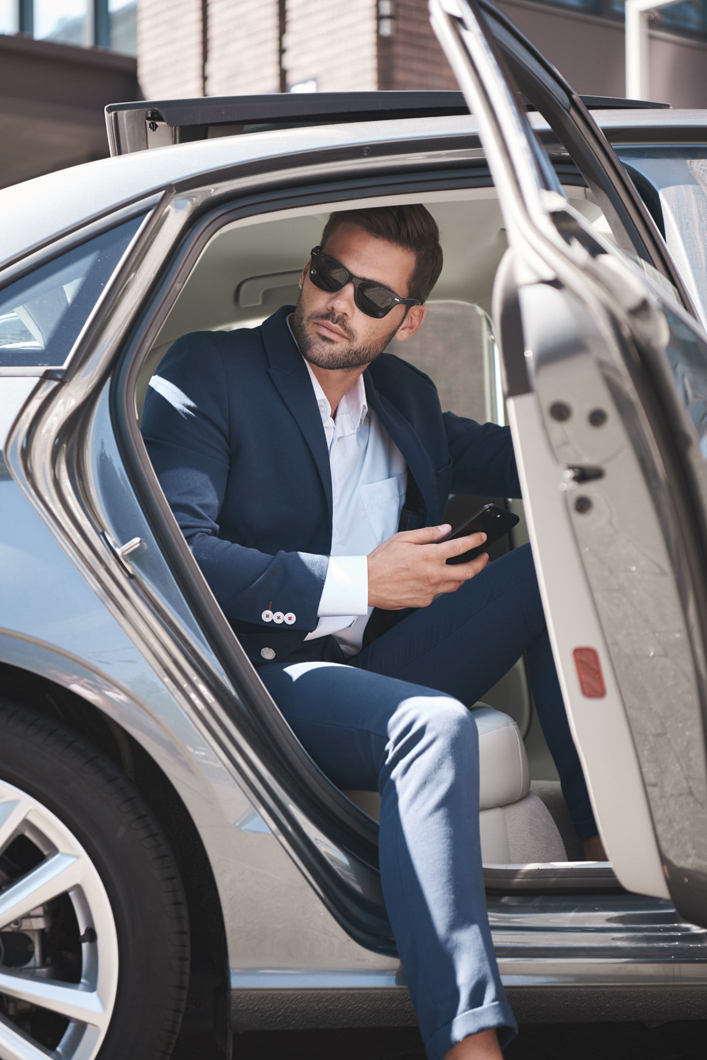Man in blue suit and sunglasses exits a car, holding a phone.
