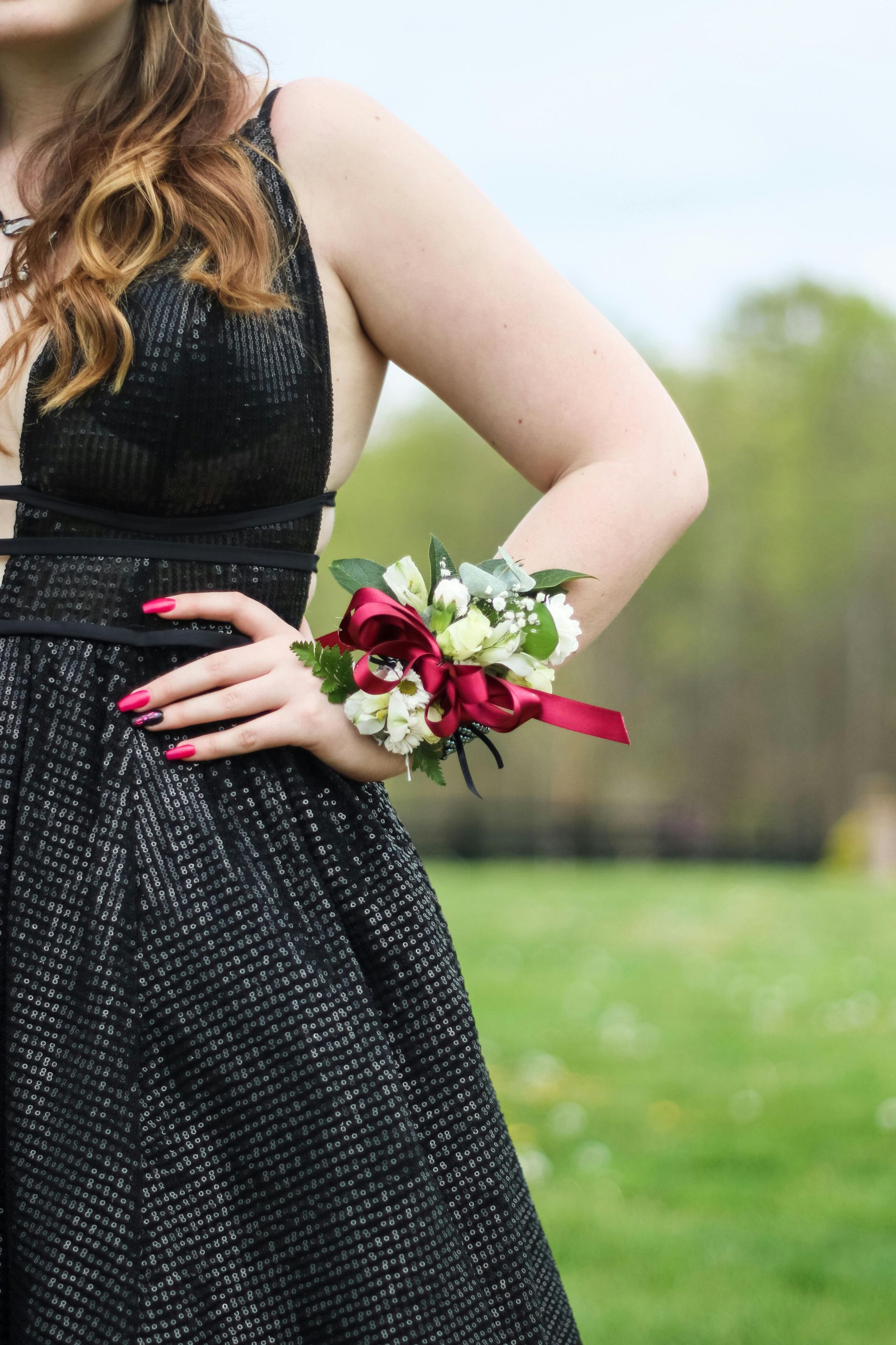 Woman in black dress with corsage on her wrist, hand on hip outdoors.