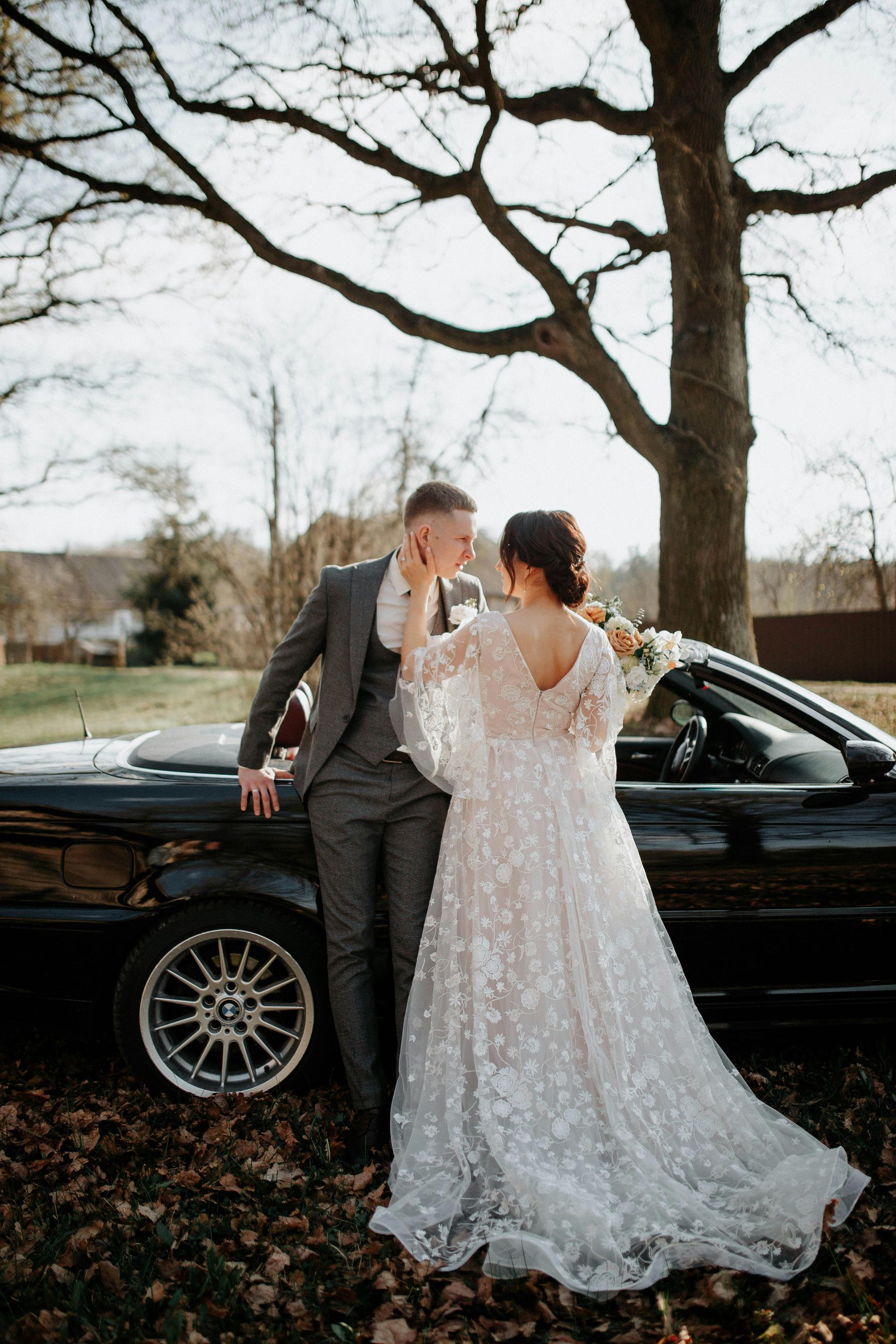 Newlyweds pose by a convertible car; the groom in gray suit, bride in a sparkly white dress.