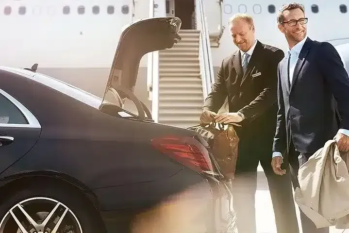 Man in suit watches as driver loads luggage into a black car near a plane.