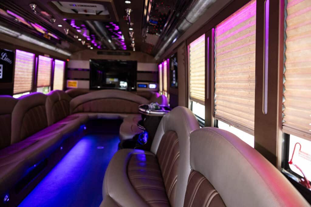 Inside of a party bus, featuring plush beige and brown seating, purple lighting, and window shades.