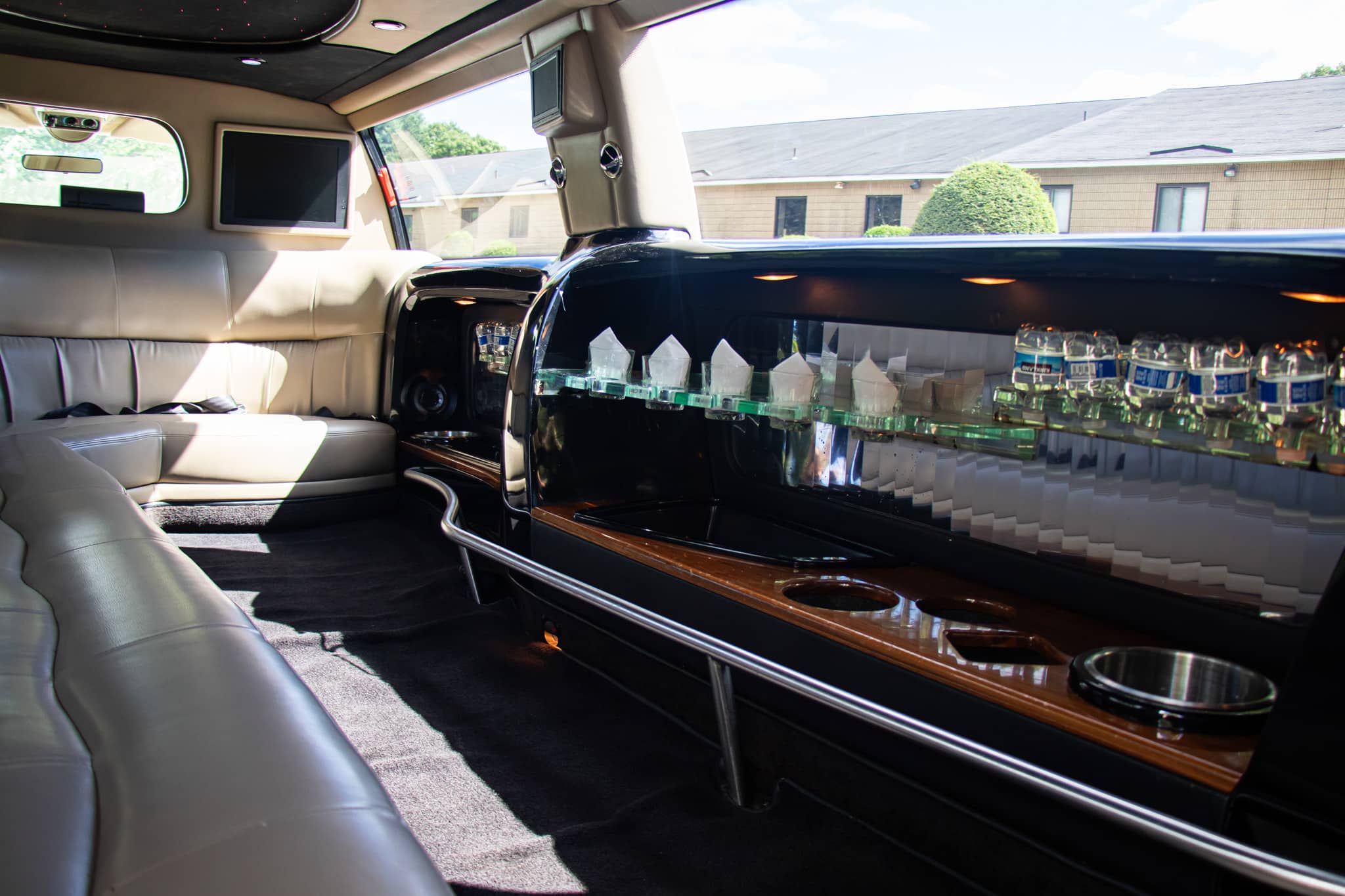 Interior of a limousine with beige leather seats, black bar, and drinks. Sunlight streams through the window.