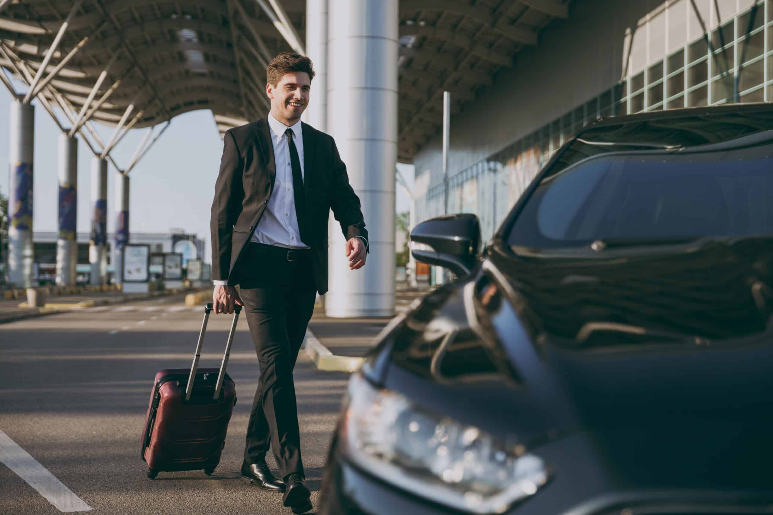 Man in a suit pulling luggage, walking towards a black car outside an airport.