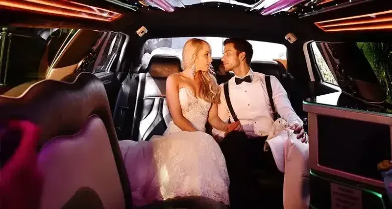 Bride and groom in wedding attire inside a limousine, holding hands, looking at each other.