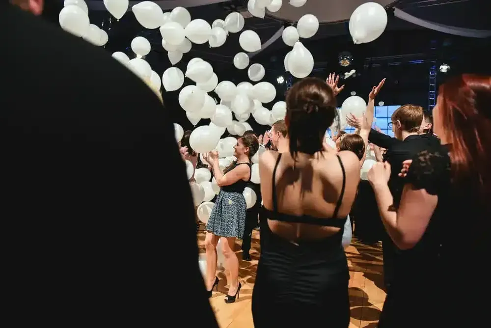 People in formal wear, celebrating with white balloons indoors.