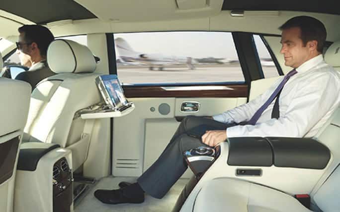 Man in a luxury car, working on a tablet. White interior, airport in the background.