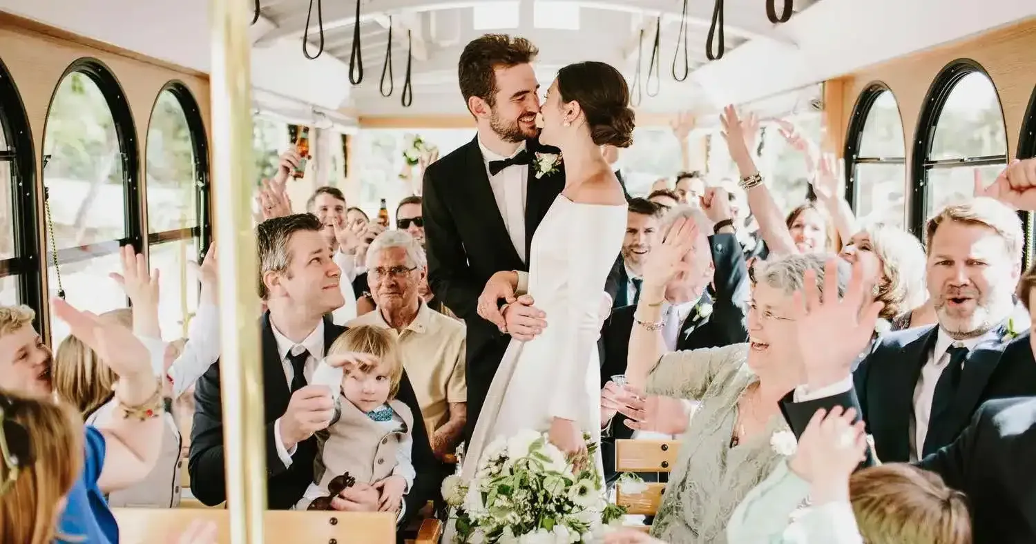Bride and groom kiss on trolley, surrounded by cheering wedding guests.
