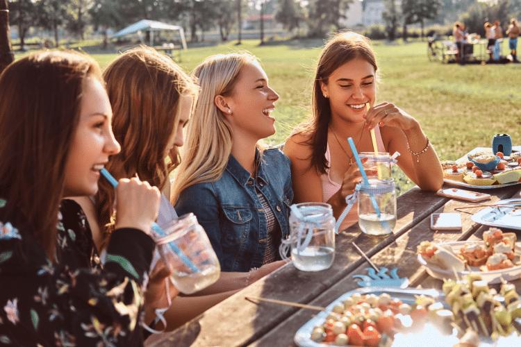 Four women laughing and enjoying drinks at a picnic table in a park on a sunny day.