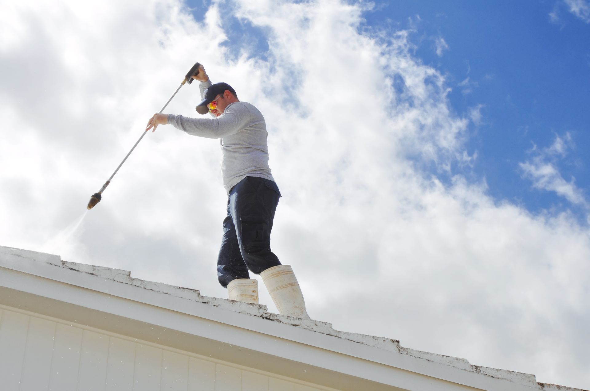 A male worker, wearing a uniform and white boots, cleans the house roof with a low-pressure washer.
