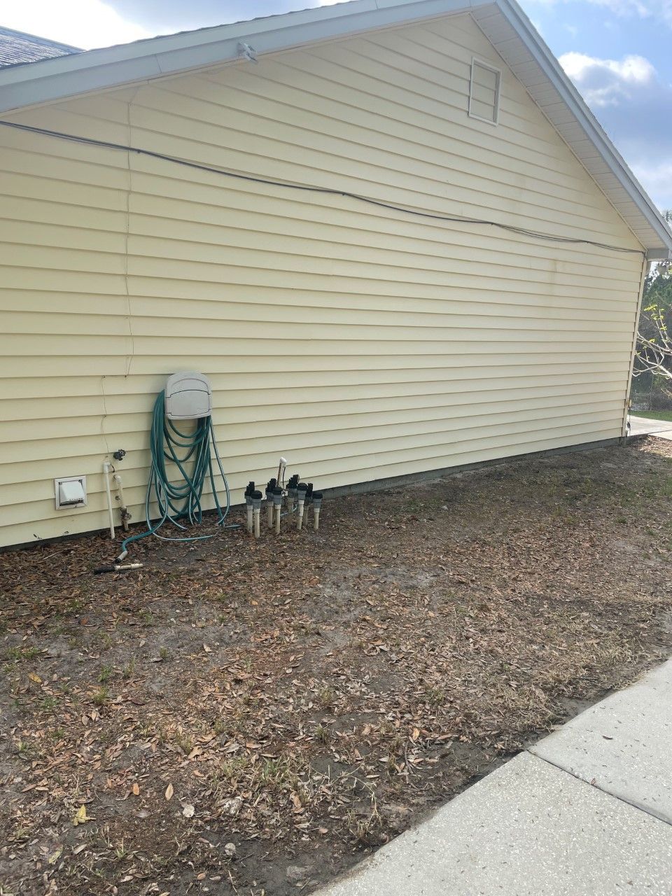 Yellow house exterior with hose, water pipes, and dry brown ground beside a sidewalk.