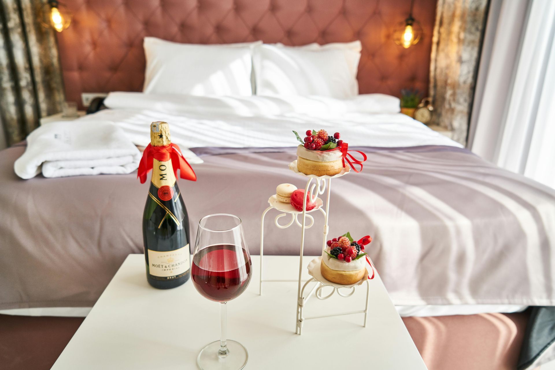A bottle of champagne , a glass of wine , and a cake on a table in front of a bed.