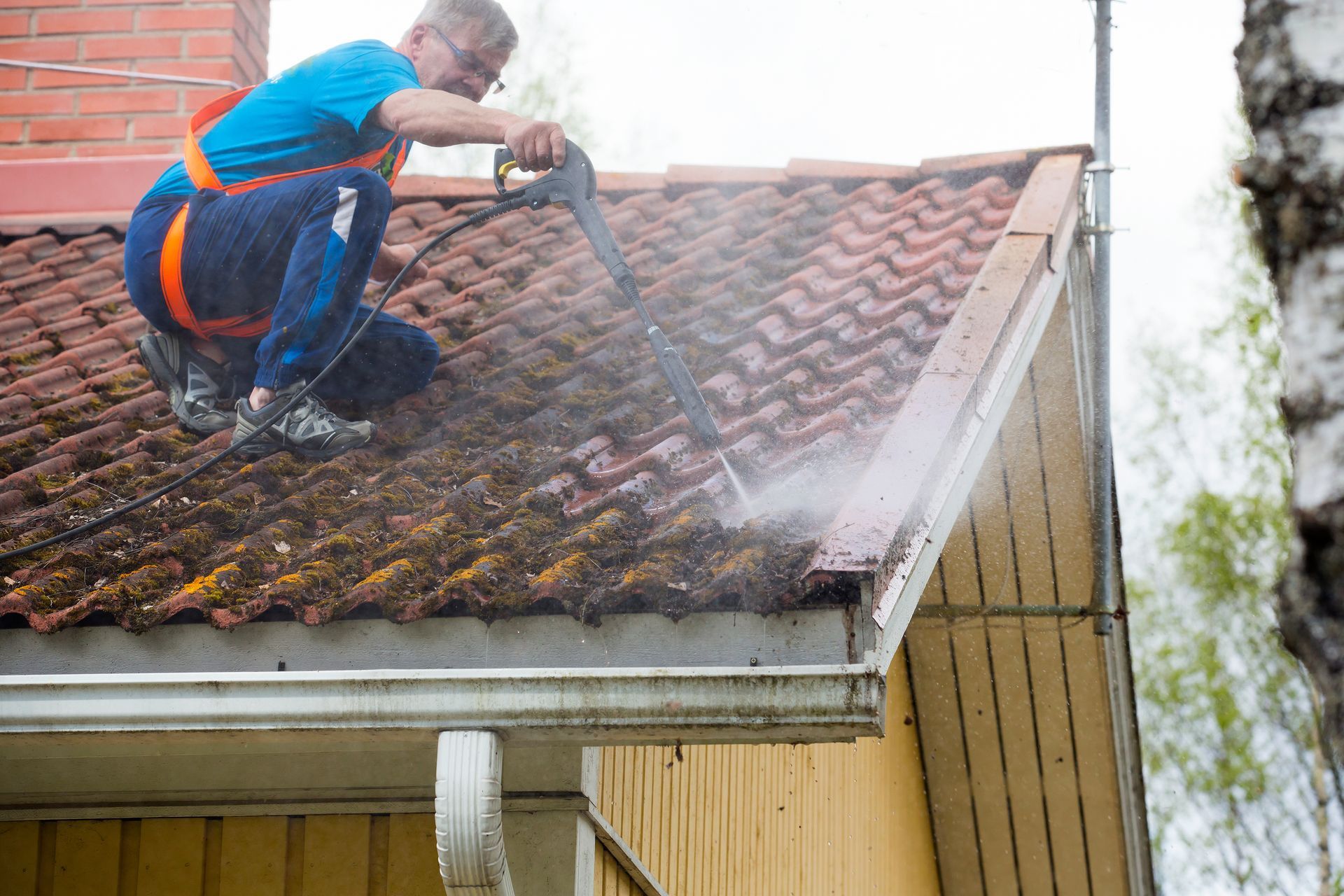 Man on a roof pressure washing red tiles, wearing safety harness, near a gutter.