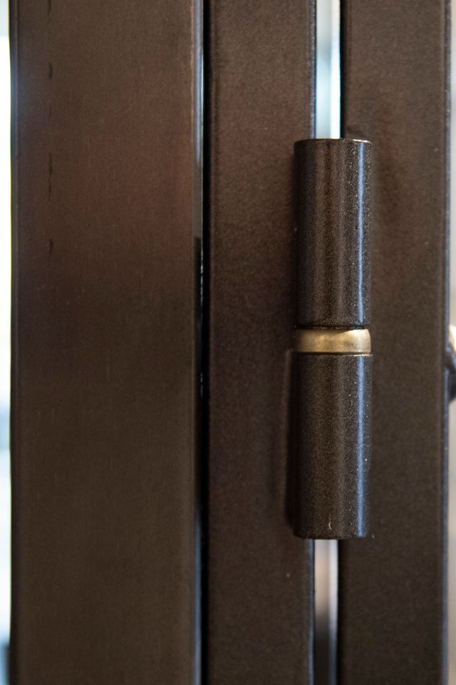 Cerniera per porta in bronzo scuro su telaio metallico, con una striscia color argento visibile sullo sfondo.