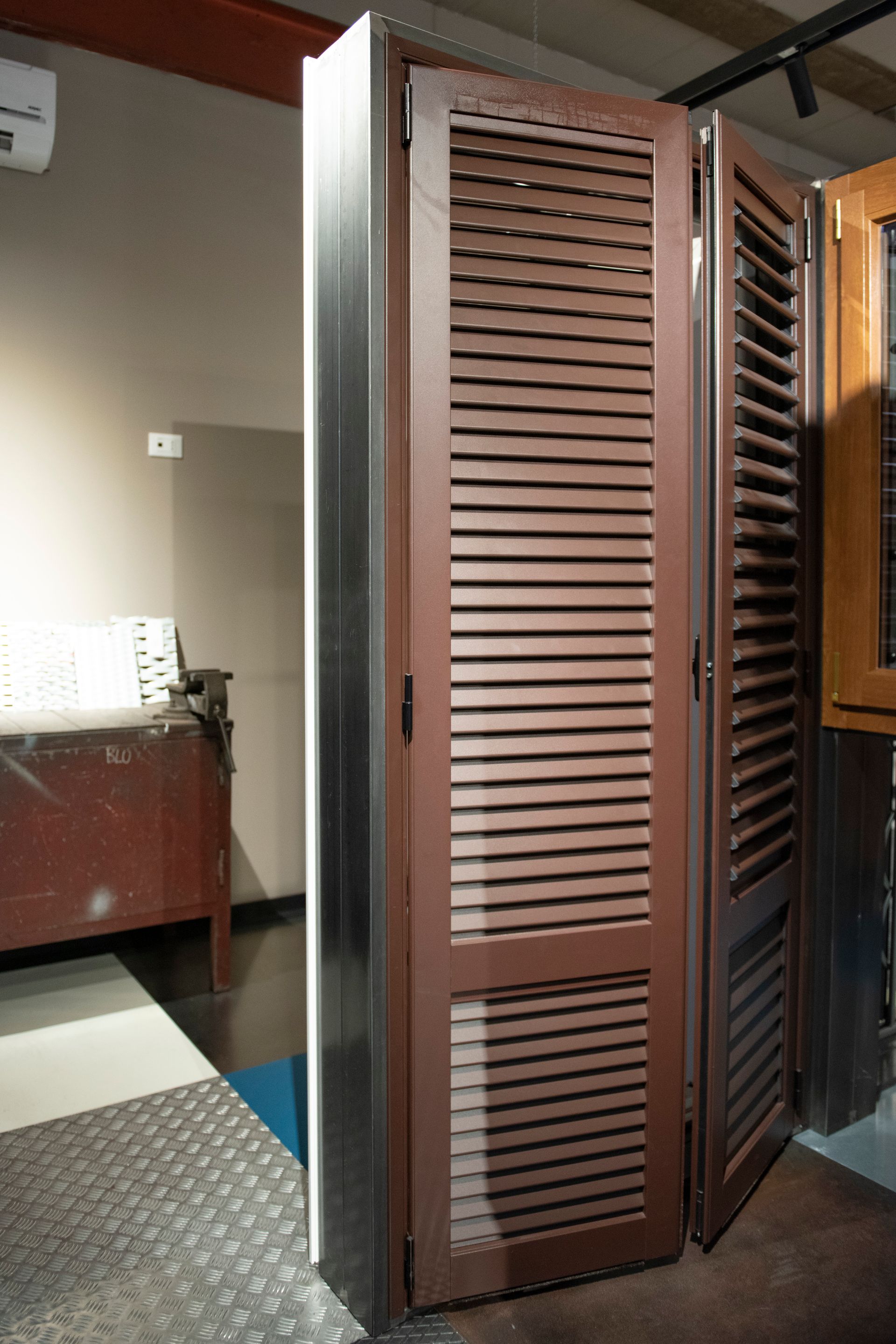 Persiane marroni con persiane, parzialmente aperte, contro una cornice metallica, in un ambiente interno ben illuminato.