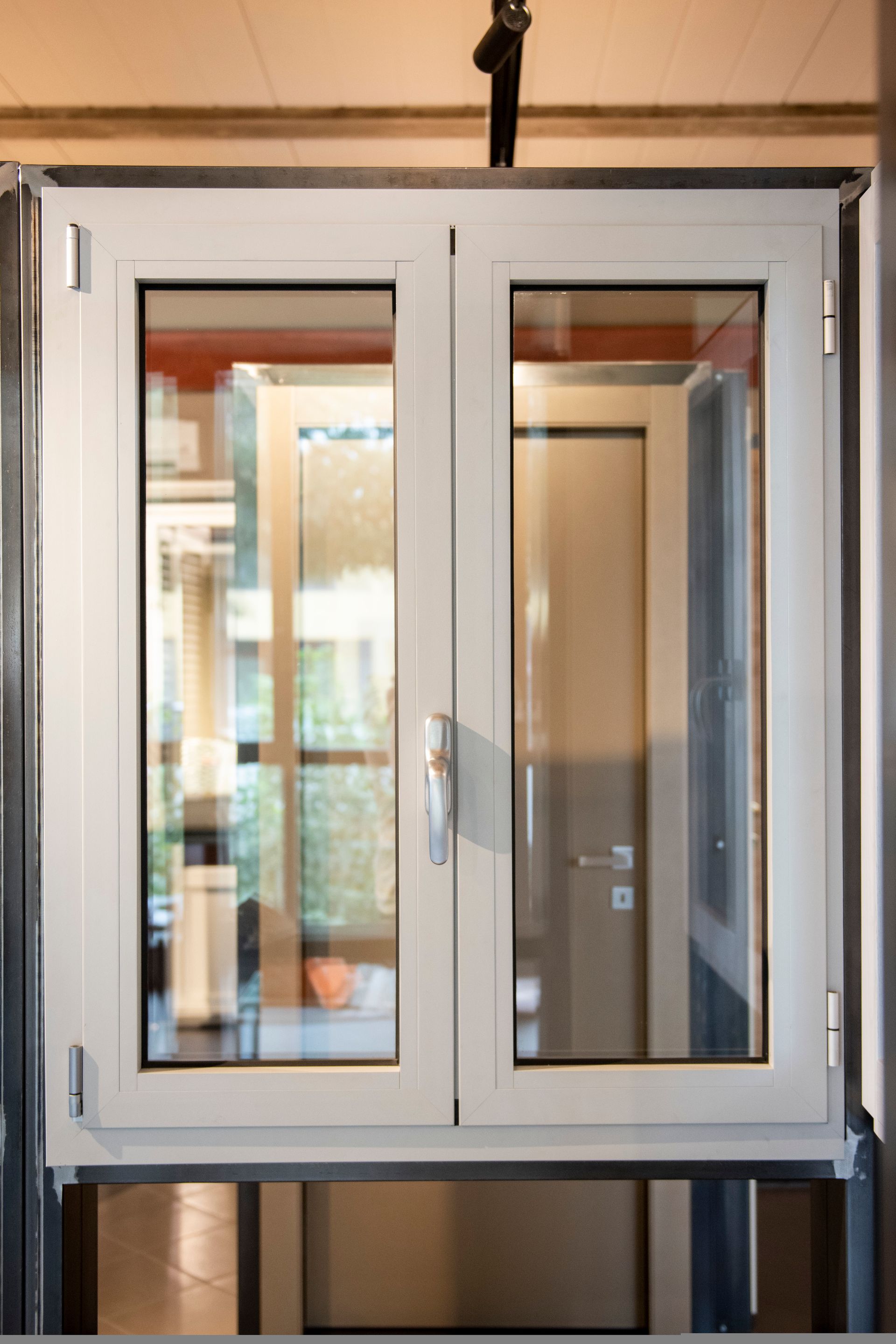 Finestra bianca con doppi vetri e maniglia, che riflette una porta interna e il verde esterno.