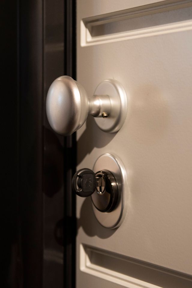 Maniglia argentata e chiave nella serratura di una porta grigio chiaro, parzialmente aperta, contro una cornice scura.