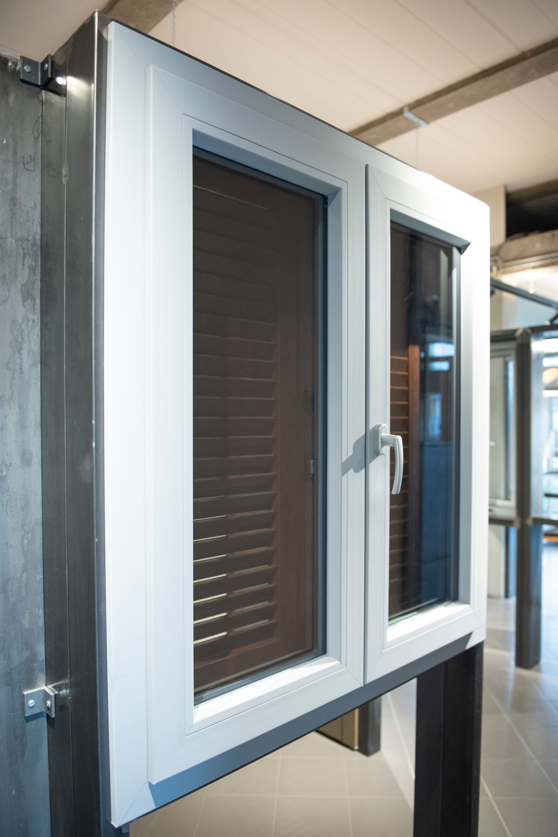 Finestra bianca a ghigliottina con maniglia, montata su un supporto scuro. La finestra è dotata di persiane e si trova in uno showroom.