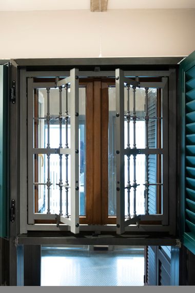 Finestra con inferriate e pannelli aperti, fiancheggiata da persiane verdi. Vista interna, luce del sole che filtra dalla finestra.