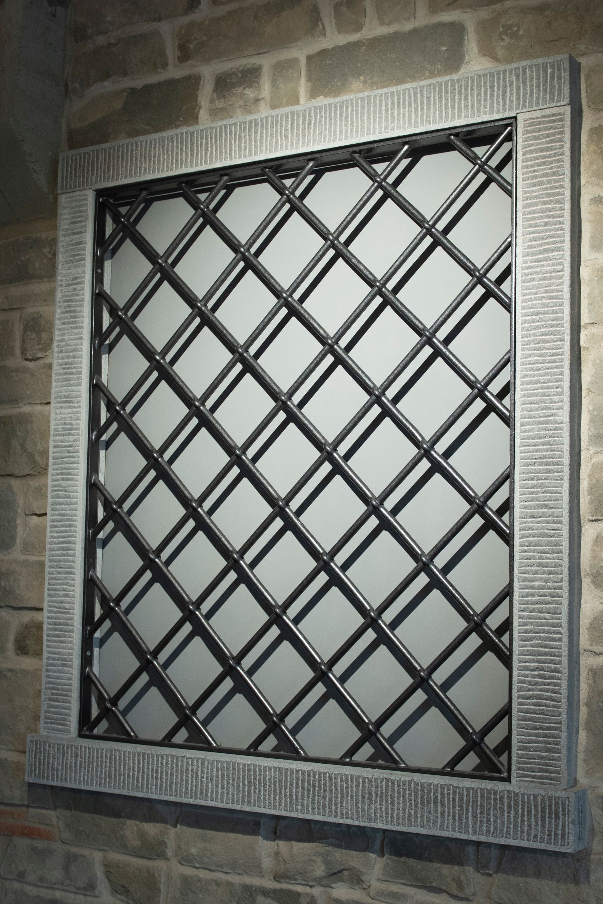 Finestra con barre metalliche a forma di diamante, incastonate in una cornice di pietra, su uno sfondo chiaro.