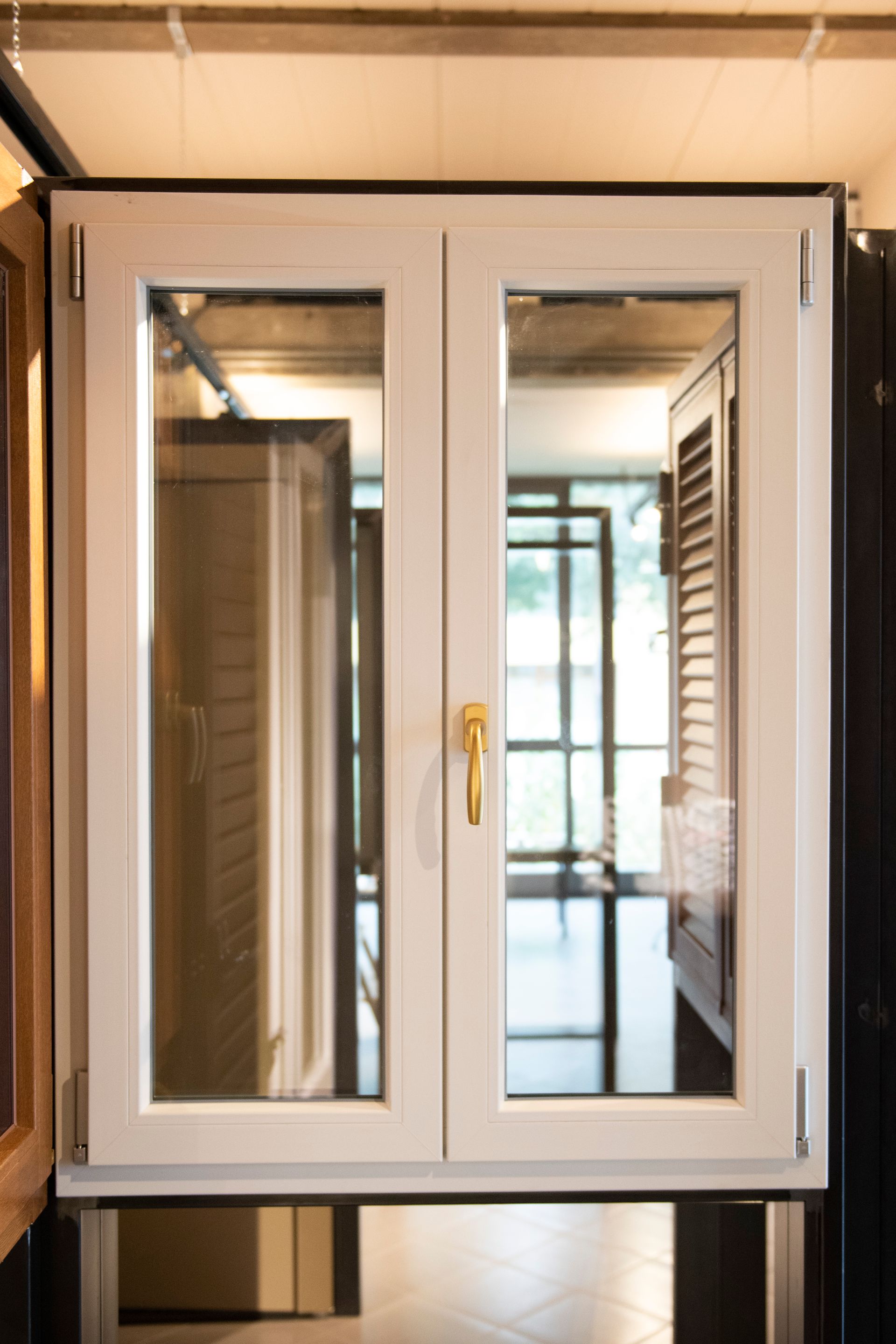 Finestra bianca a ghigliottina con maniglia dorata. Persiane aperte e riflesso di una stanza visibile attraverso il vetro.