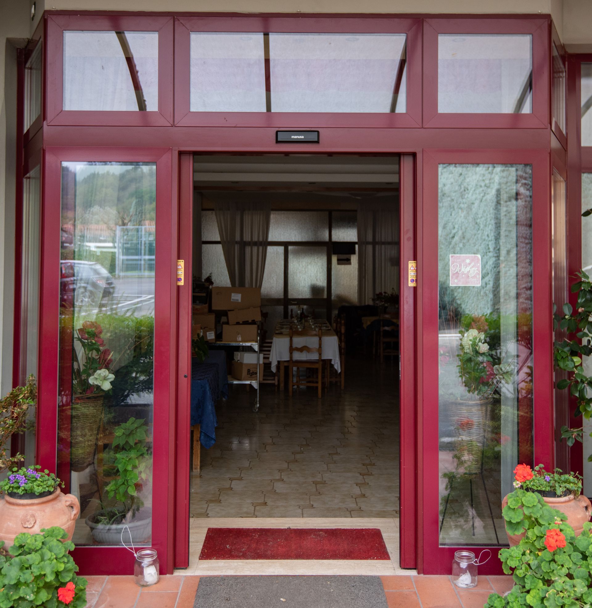 Un portale con cornice rossa conduce a un ristorante. L'esterno è decorato con piante in vaso, finestre in vetro e uno zerbino rosso.