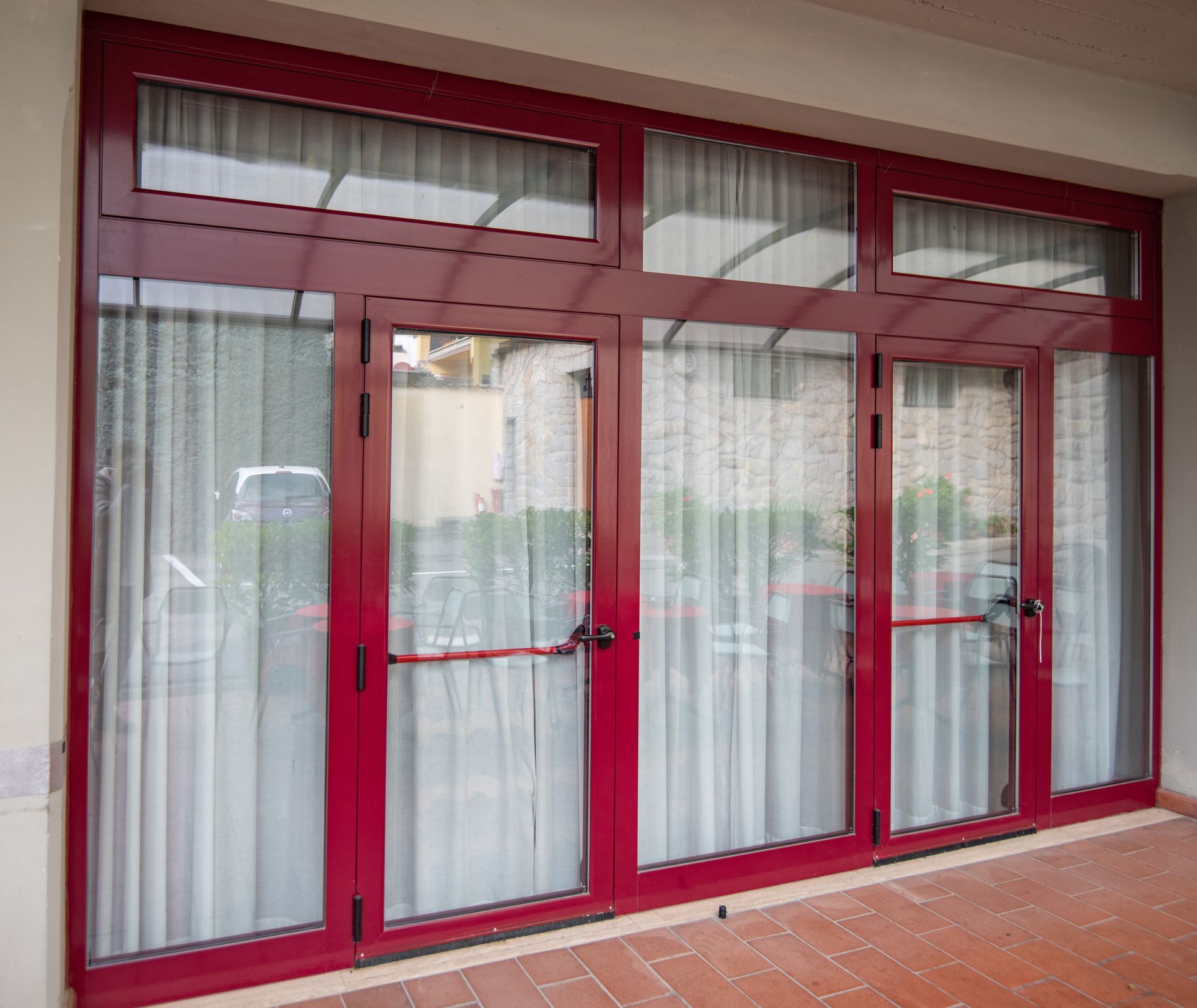 Porte in vetro con cornice rossa, tende bianche, una pensilina e una barra rossa per le uscite di emergenza.