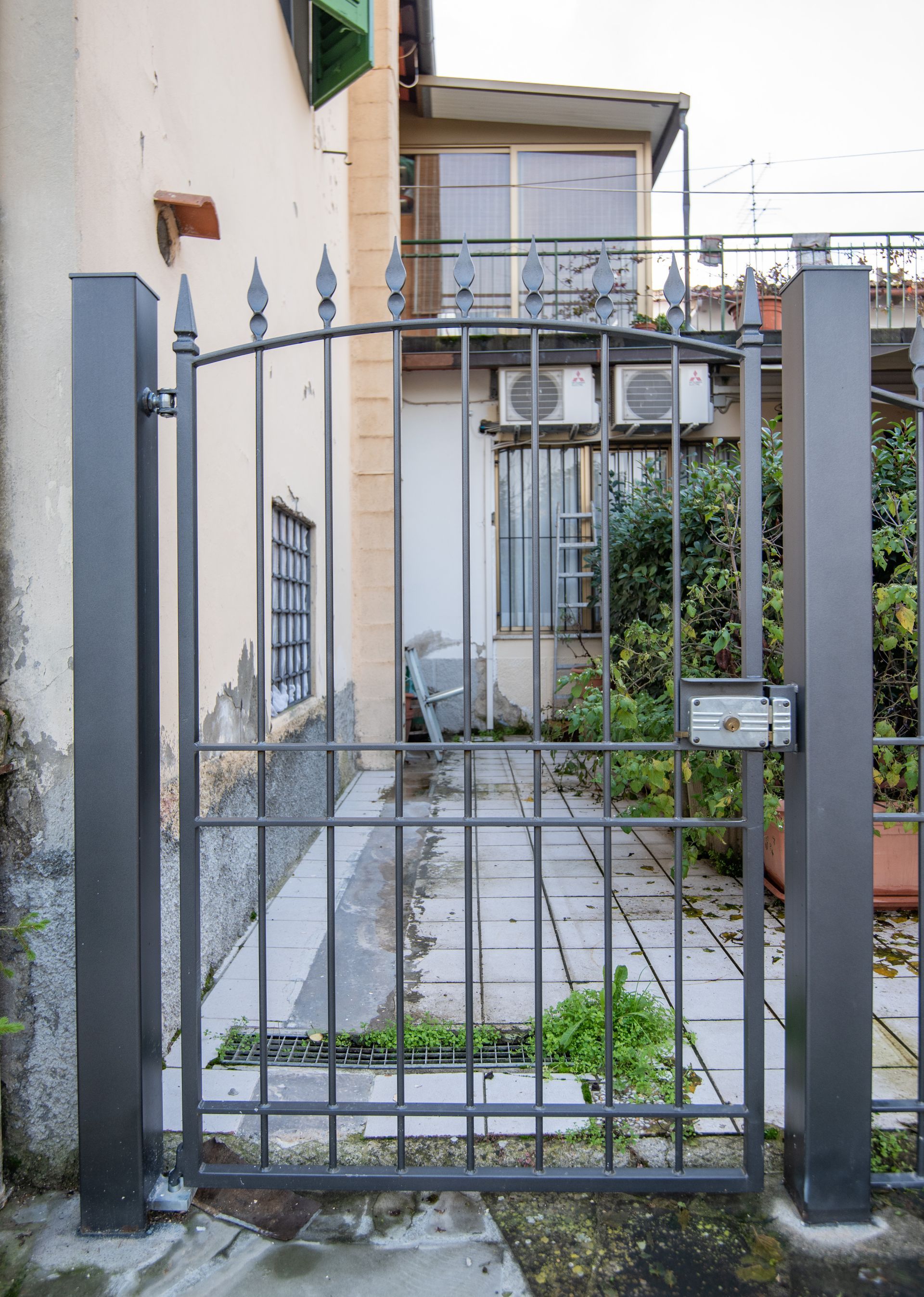 Cancello metallico grigio scuro, chiuso a chiave, incastonato nel cemento. Conduce a un cortile con un edificio sullo sfondo.