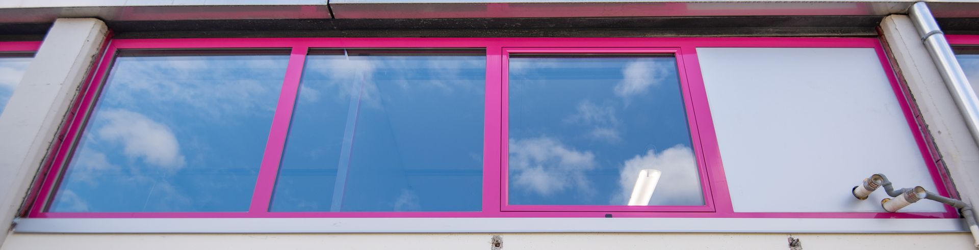 Finestre con cornici rosa che riflettono un cielo azzurro con nuvole. Un pannello bianco si trova sulla destra.