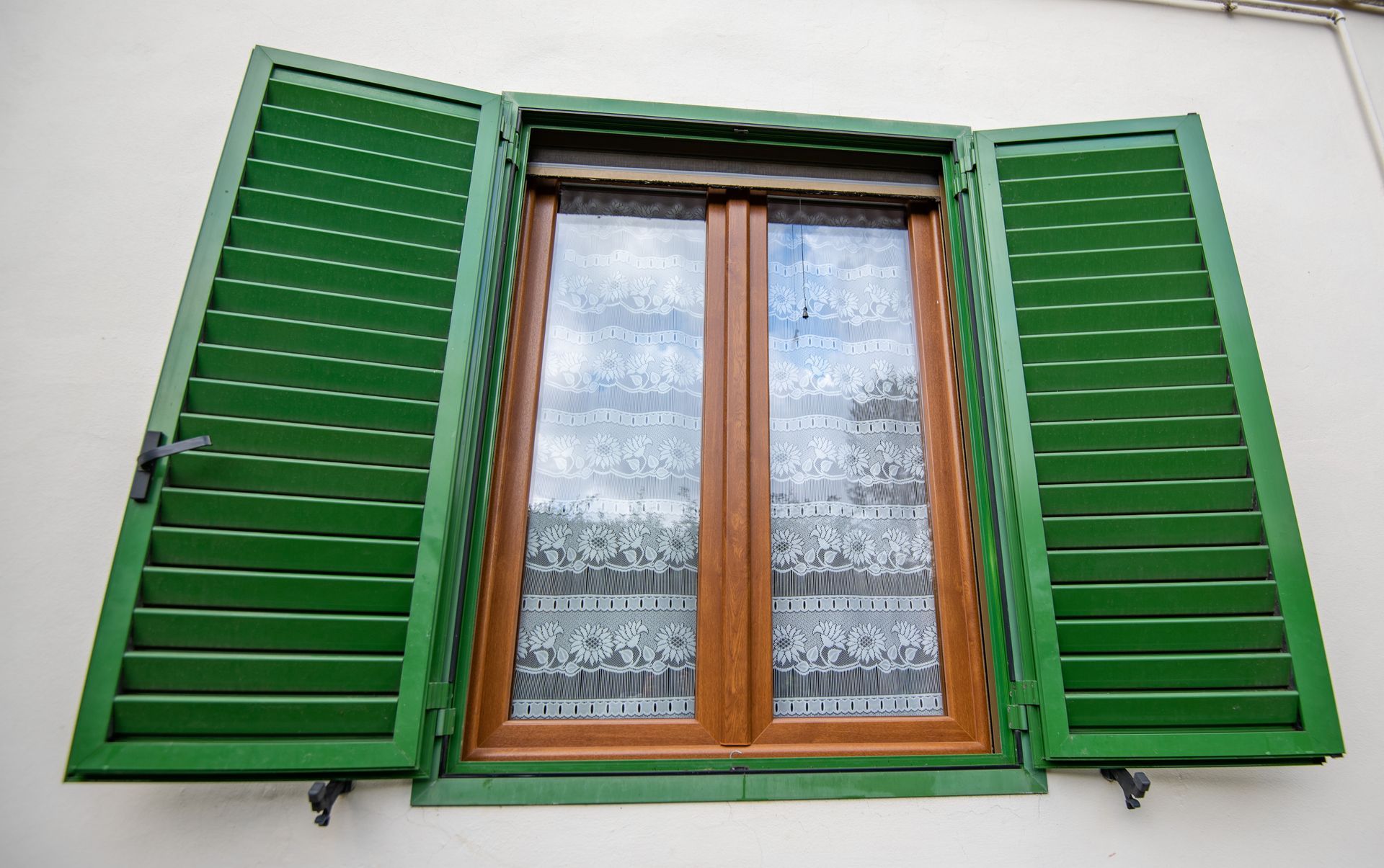 Persiane verdi che incorniciano una finestra con finiture marroni, tende di pizzo.
