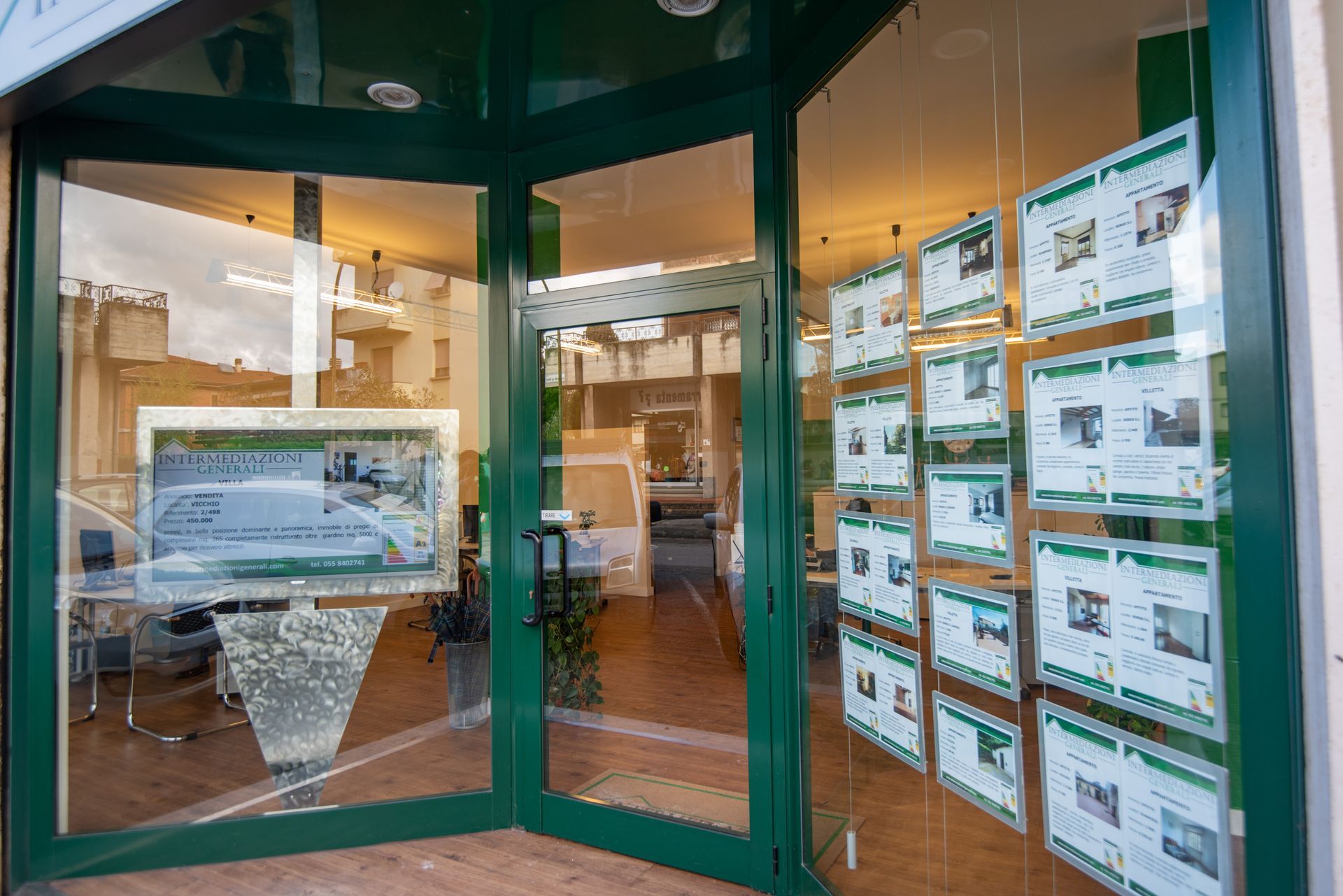 Ufficio immobiliare con struttura verde, annunci immobiliari alle finestre, porte in vetro e uno schermo all'interno.