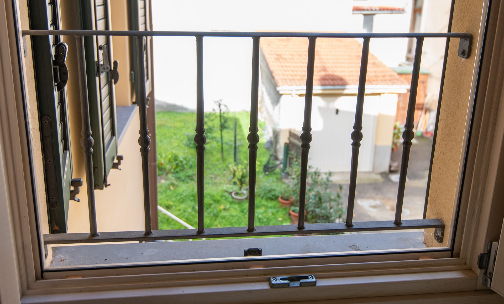 Finestra con inferriate decorative in metallo che si affaccia su un cortile verde e su un edificio con tetto in tegole rosse.