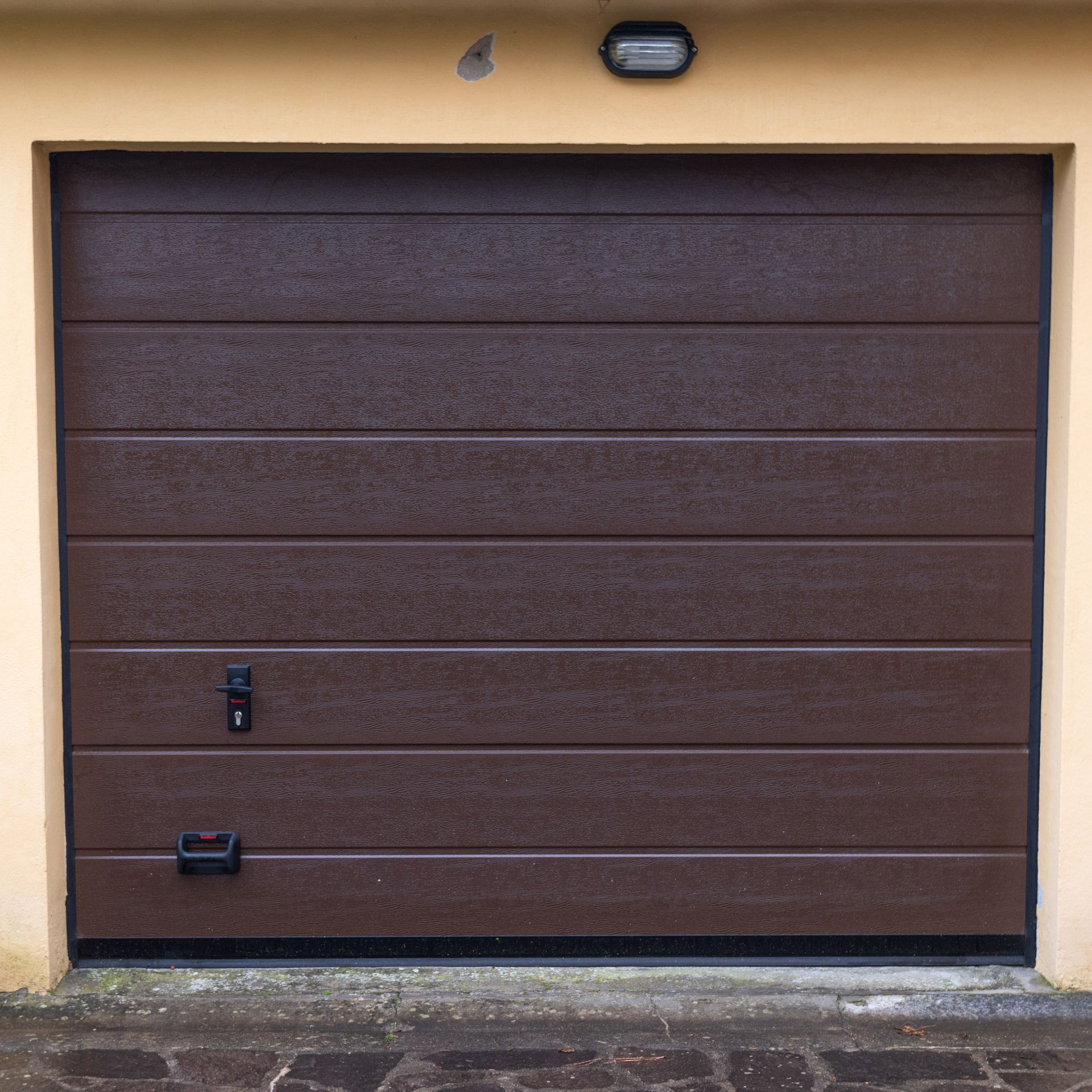 Porta da garage marrone con pannelli orizzontali; ferramenta nera. Rifiniture e parete beige.