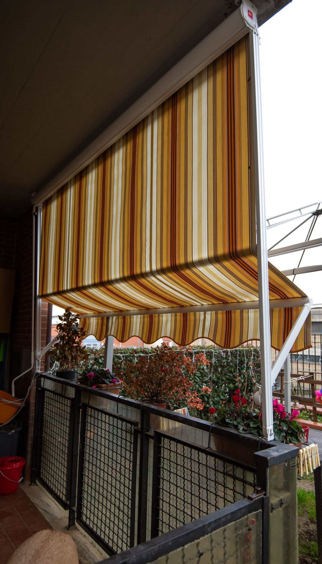 Tenda da sole a strisce beige e marroni estesa su un balcone con ringhiera e piante in vaso.