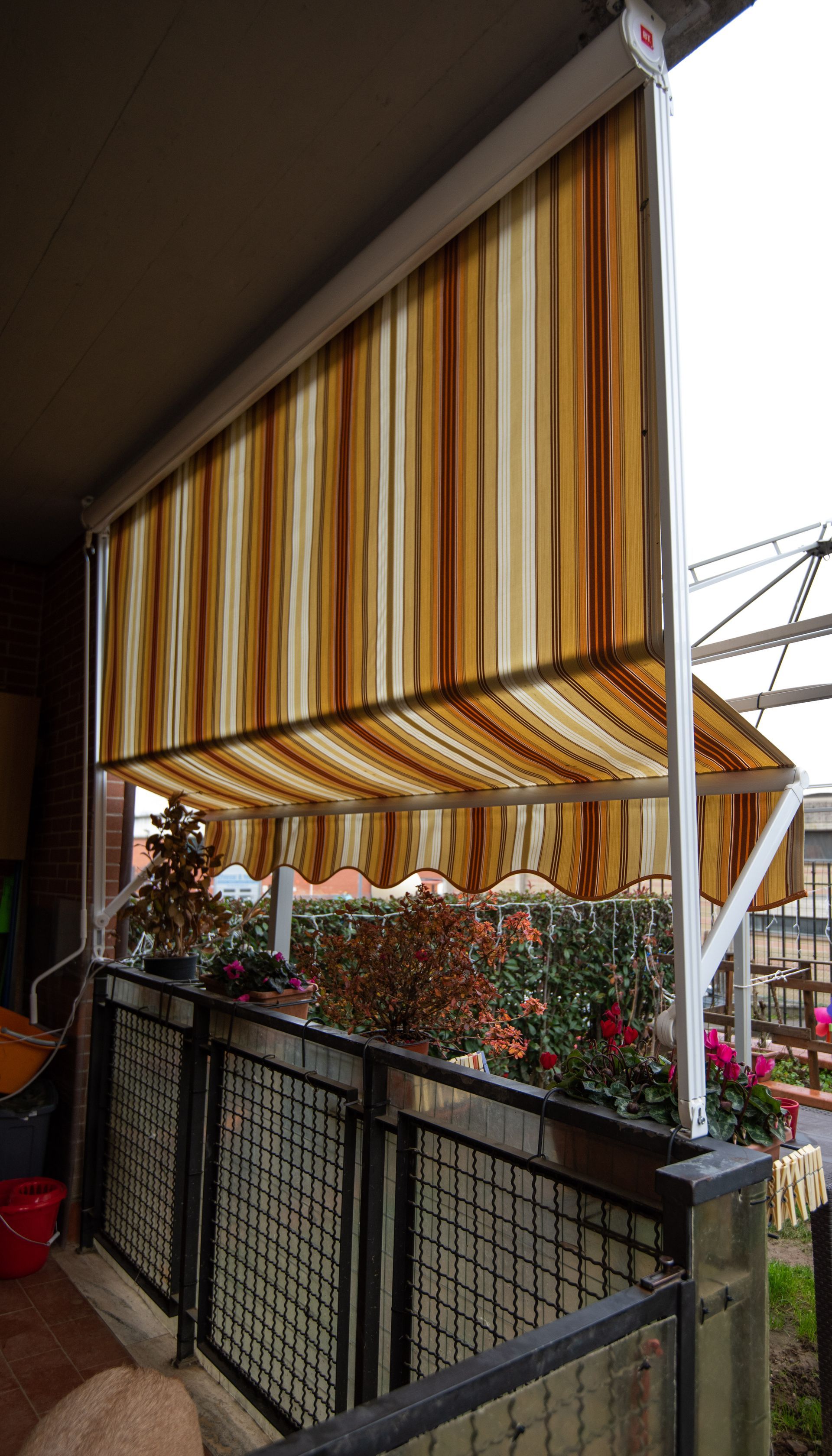 Tenda da sole a strisce beige e marroni estesa su un balcone con ringhiera e piante in vaso.