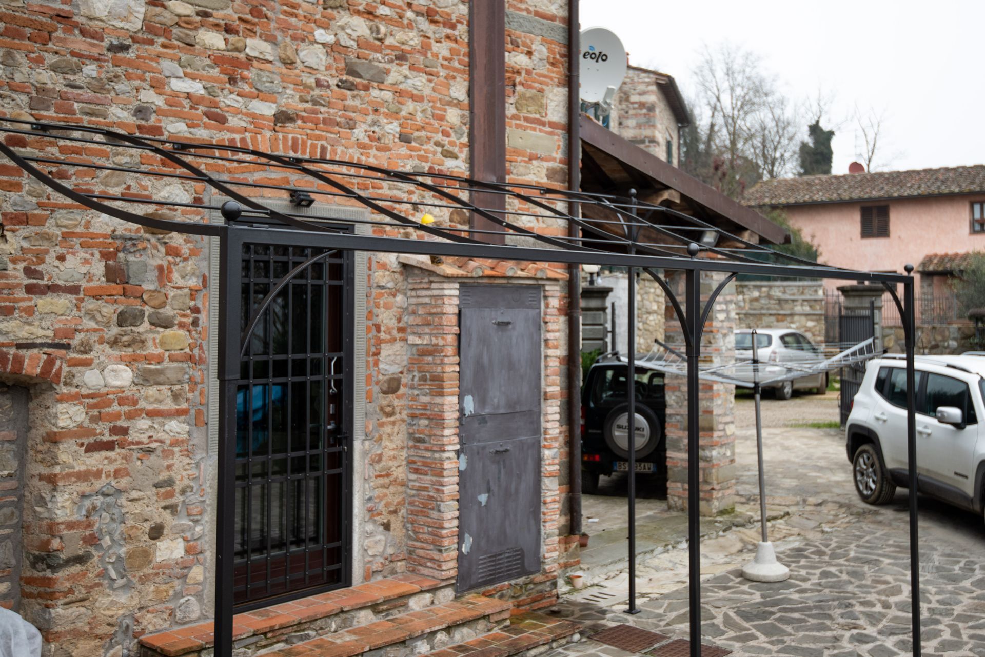 Pergola in metallo nero all'esterno di un edificio in mattoni con cancello nero e porta in legno. Le auto sono parcheggiate nelle vicinanze.
