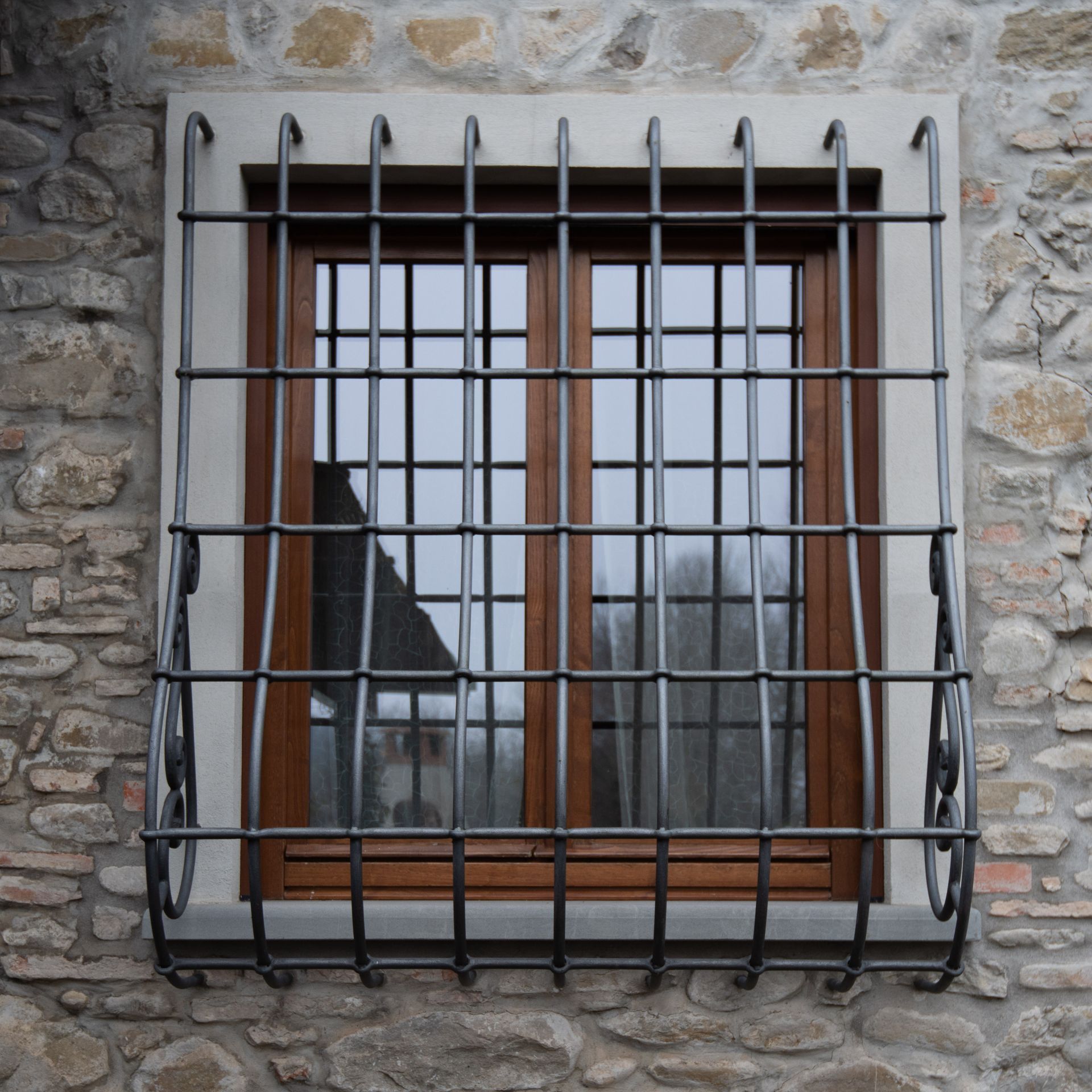 Finestra con sbarre di metallo su un muro di pietra, cornice marrone, in cui si riflettono una casa e degli alberi.
