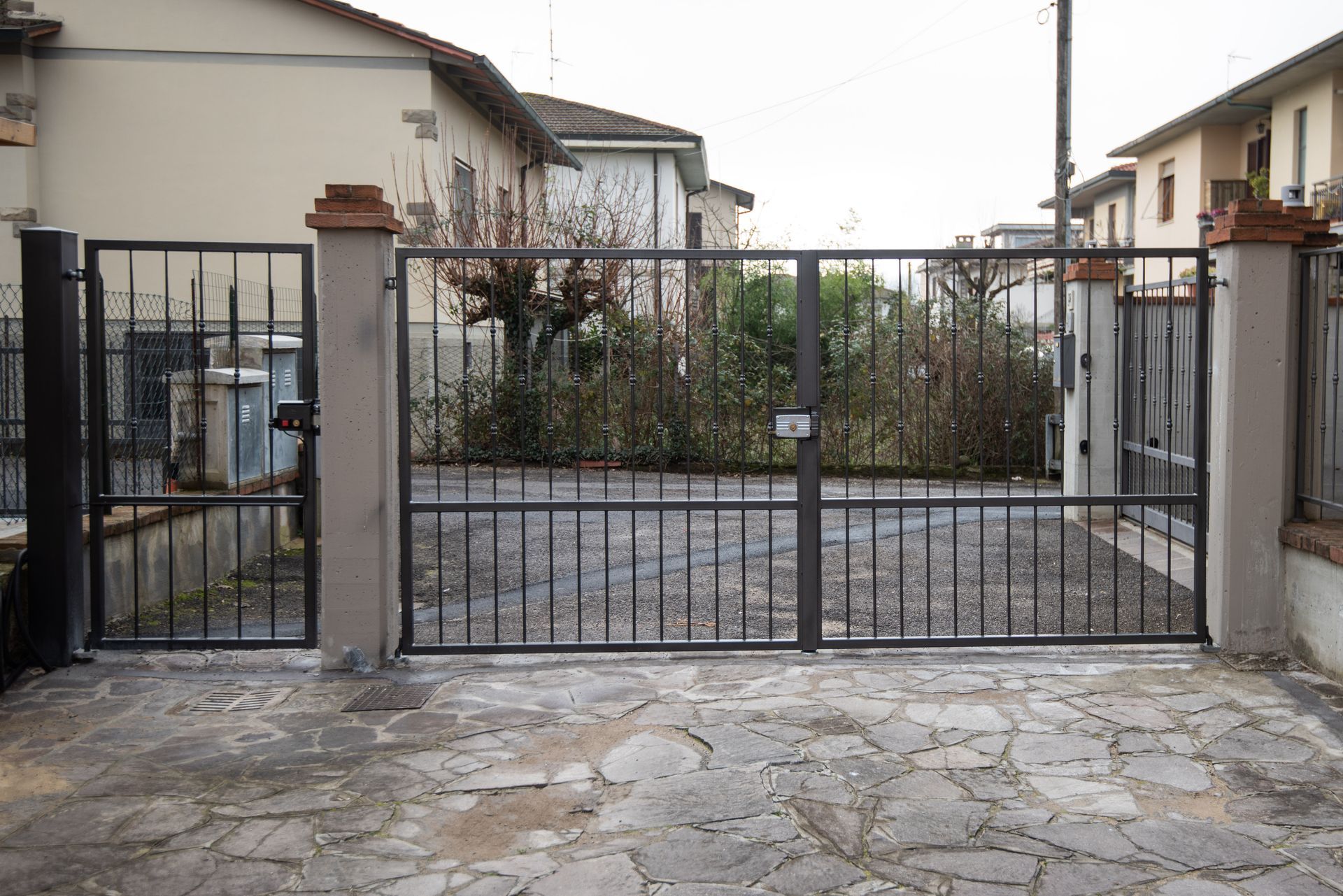 Cancello e recinzione in metallo, di fronte a un vialetto in pietra e a edifici residenziali.