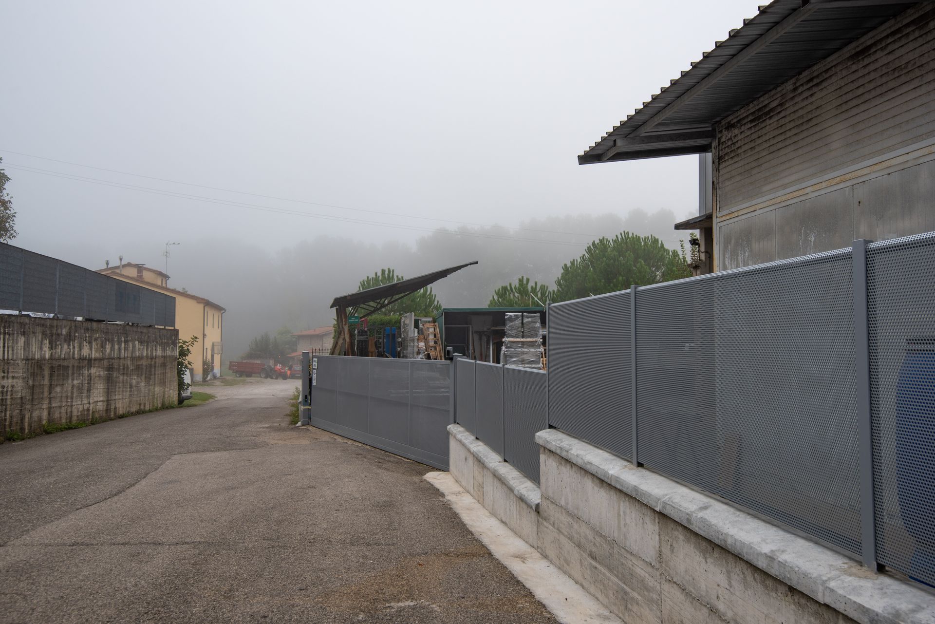 Scena di strada nebbiosa con edifici e recinzioni metalliche.
